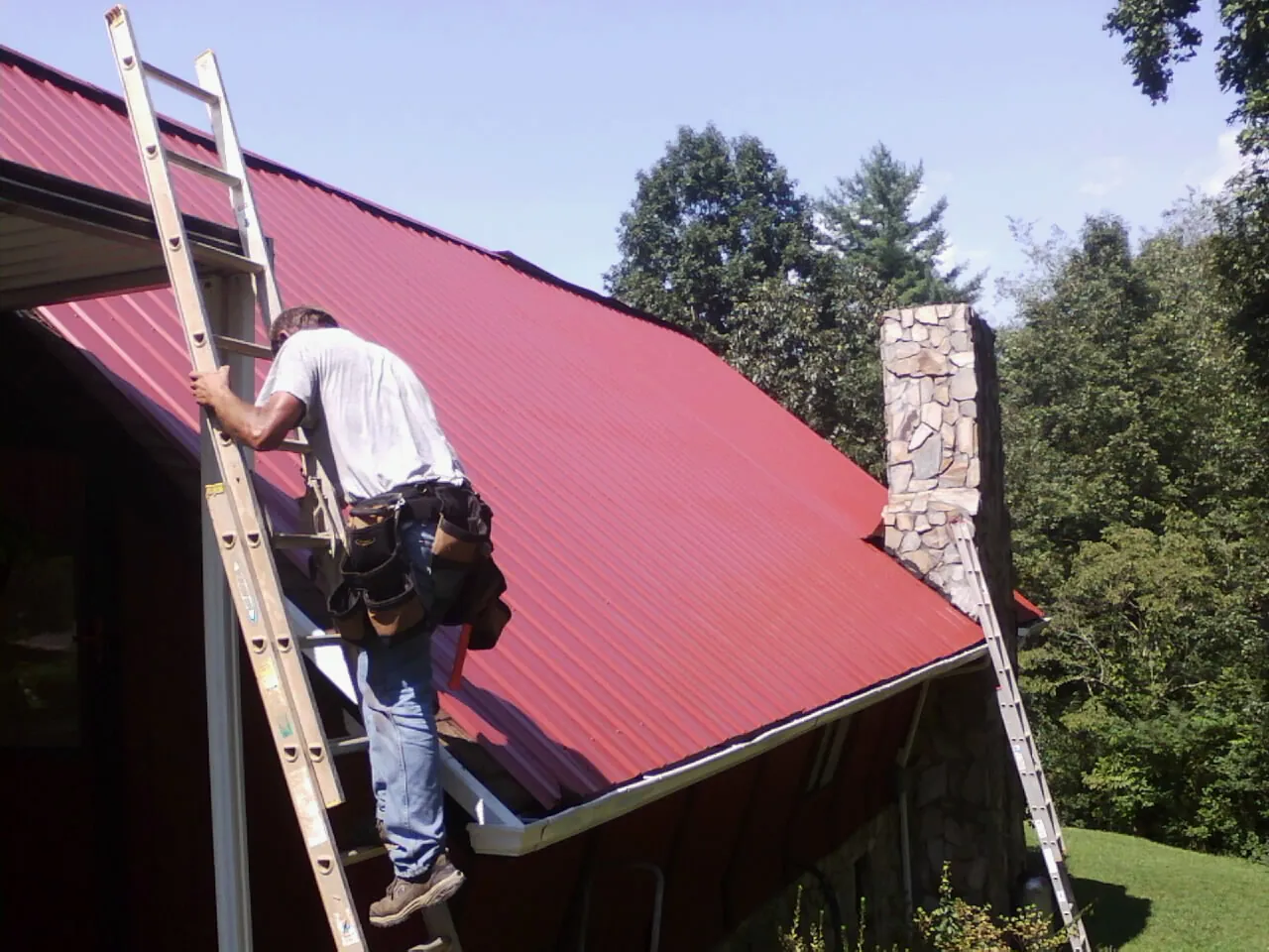 Roofer on a ladder beside a red metal roof and stone chimney — Soffit & Fascia Repair by Roofing Brothers in Fulton